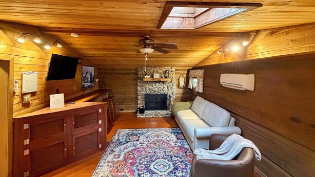 A log cabin interior with a stone fireplace, wooden ceiling, a sofa, a television, and a desk with a computer.