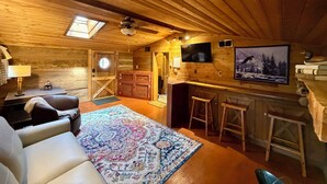 A cozy cabin interior with a wooden ceiling, a large rug, a sofa, a desk, and a mounted TV.