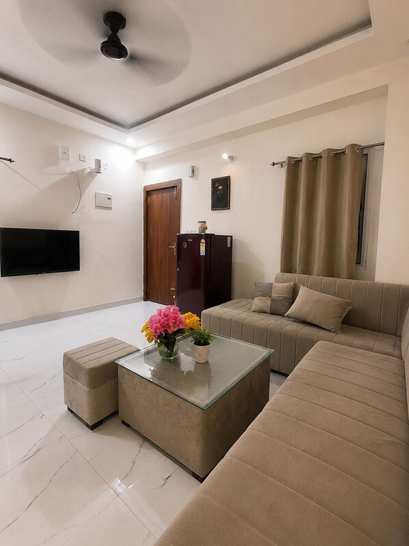 A living room with a sofa, a glass coffee table, a television, a ceiling fan, and a door. - Cozy 1 BHK Staycation | Patio | Tapovan, Rishikesh (Rishikesh)
