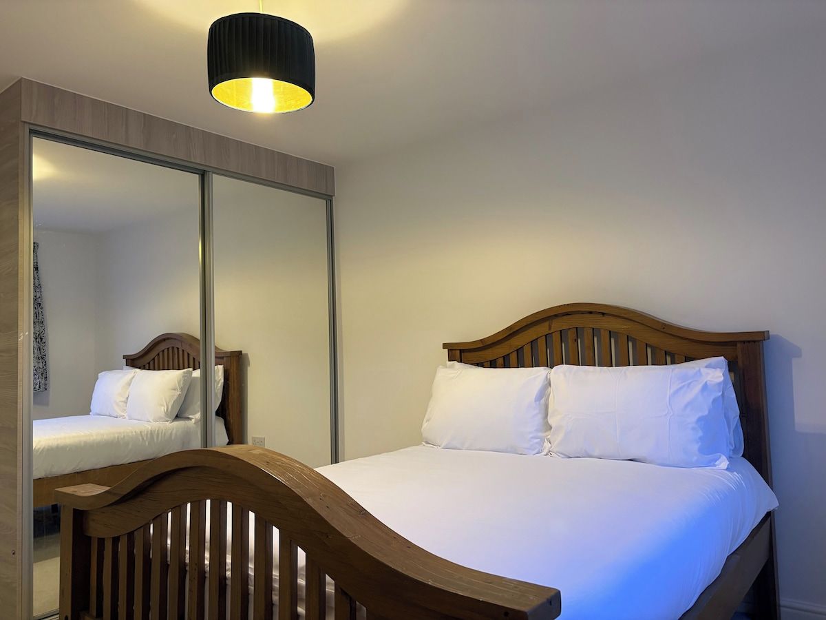 A bedroom with a wooden bed, white bedding, a mirror, and a ceiling light fixture.