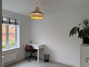 Une pièce avec un bureau blanc, une chaise, une lampe suspendue, un radiateur, une fenêtre avec des stores et une plante en pot.