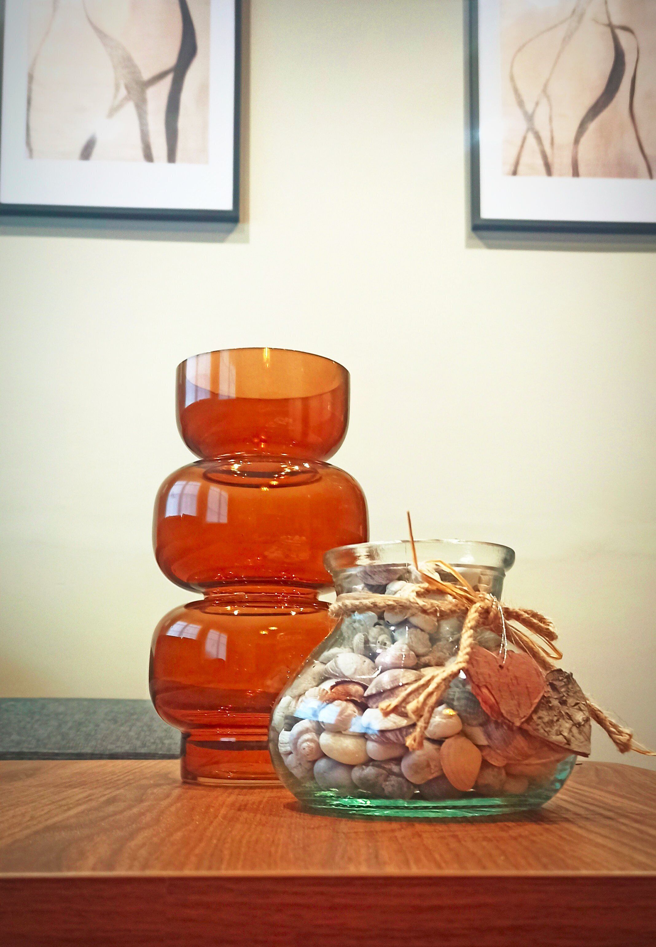 A stack of orange glass vases and a glass jar filled with pebbles and twine, placed on a wooden surface.