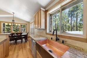 Eine Küche mit Holzausstattung, einem Kupferspülbecken, einem Essbereich mit Tisch und Stühlen sowie großen Fenstern mit Blick auf einen Wald.