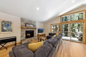 A living room with a stone fireplace, a piano, a large sofa, and a view of the outdoors through large windows. - Family Delight at Tahoe (South Lake Tahoe)