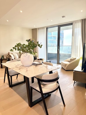 A modern living room with a marble dining table, plush chairs, a large potted plant, and a city view through the glass door. - Cosy 1 bedroom luxe apartment in the heart of London (London)