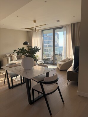 Een moderne woonkamer met een eetgedeelte, een groot raam met gordijnen, een plafondventilator en uitzicht op een gebouw.