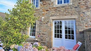 A stone building with a large window and a door, surrounded by lush greenery and flowers. - Coastal Stone House in Lannilis for 5 (Lannilis)