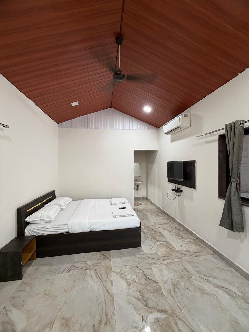 A bedroom with a bed, a ceiling fan, an air conditioning unit, a television, and a bathroom visible in the background.