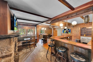Um interior rústico com vigas de madeira, bancadas de pedra, banquetas de bar e uma TV fixada na parede.