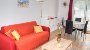 Un salon avec un canapé rouge, une petite table sur laquelle est posé un vase, un coin repas avec des chaises et une fenêtre avec des stores.