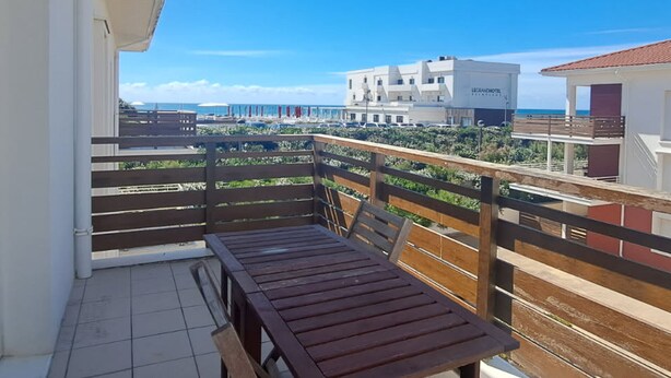 Een balkon met een houten tafel en stoelen, met uitzicht op een strand en hotelgebouwen.
