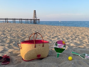 Eine Strandansicht mit einer Strohbag, einem Strandball und einem Paddel im Sand.
