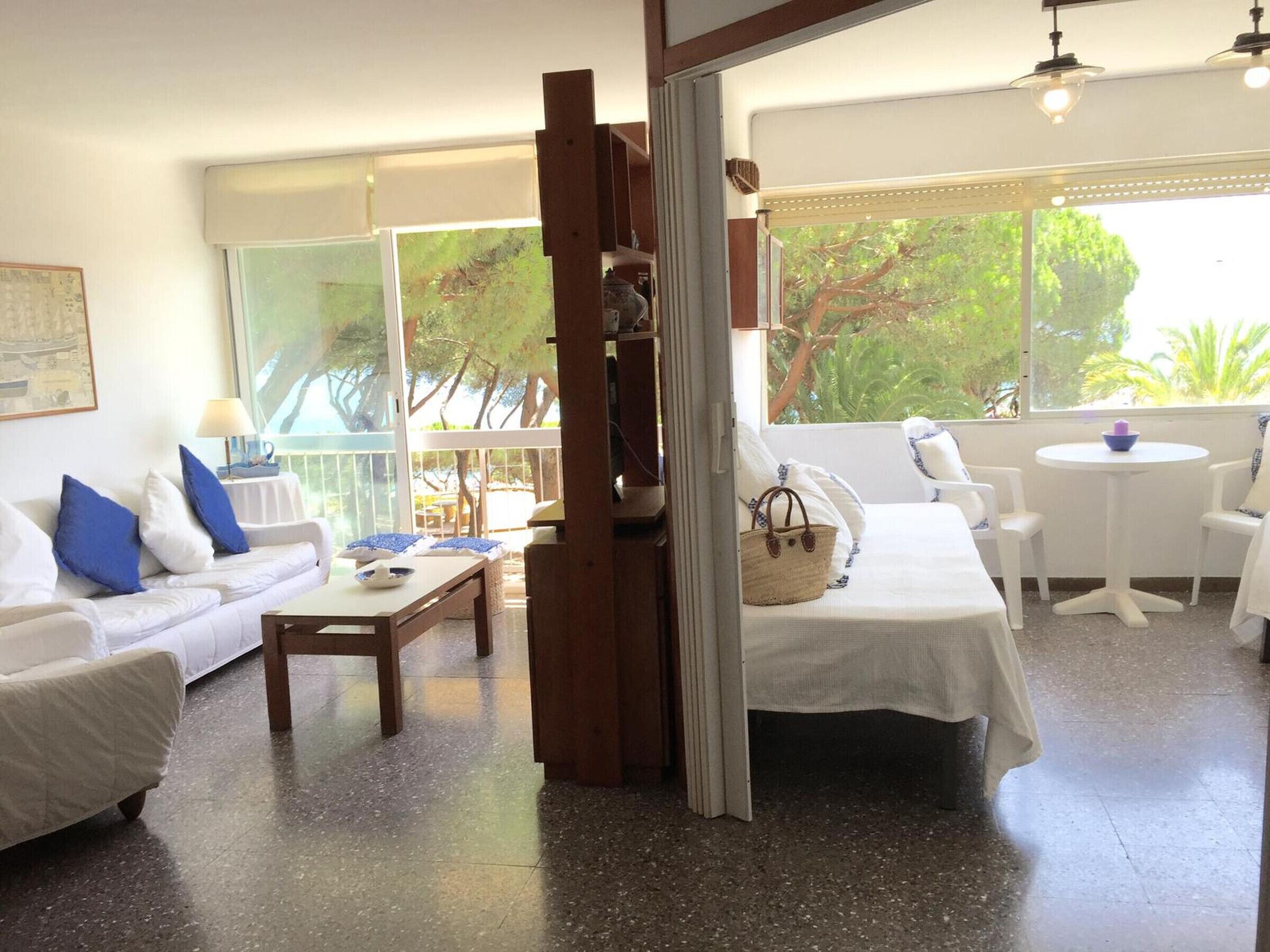 A bright room with a white sofa, a wooden table, and a balcony with a view of trees.