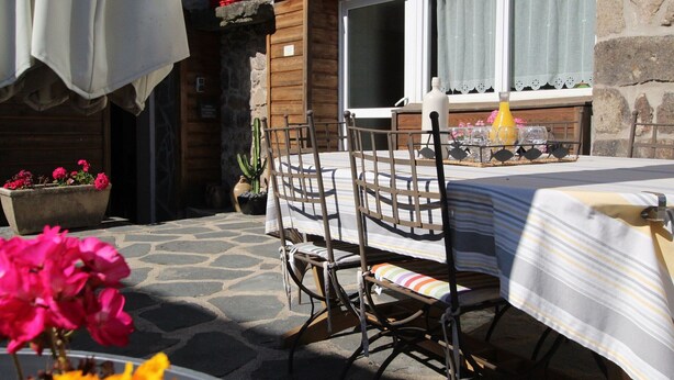 An outdoor patio with a table and chairs, a potted plant, and a window with curtains.