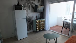 Ein Zimmer mit Kühlschrank, Fernseher auf einem Schrank, einem kleinen Tisch mit Stühlen und einem Balkon mit Aussicht.