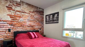Quarto com parede de tijolos, cama com colcha vermelha, piso xadrez em preto e branco e janela com vista para o exterior.