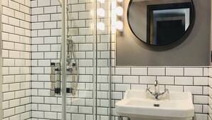 Um banheiro com azulejos brancos tipo metrô, um chuveiro com box de vidro, uma pia branca com detalhes cromados e um espelho redondo com iluminação superior.