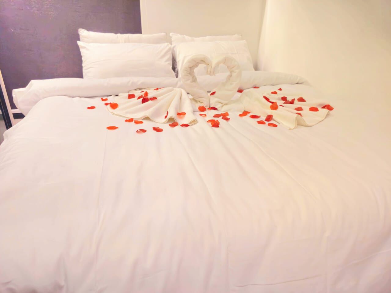 Una cama con sábanas blancas, un cojín con forma de corazón y pétalos de rosa esparcidos.