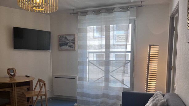 A living room with a sofa, a table, a TV mounted on the wall, a radiator, and a window with a curtain. - Quiet apartment with parking - 4 guests (Cauterets)