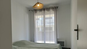 A room with two beds, a window with sheer curtains, a hanging light fixture, and a radiator. - Modern apartment with parking - 4 guests (Le Lavandou)