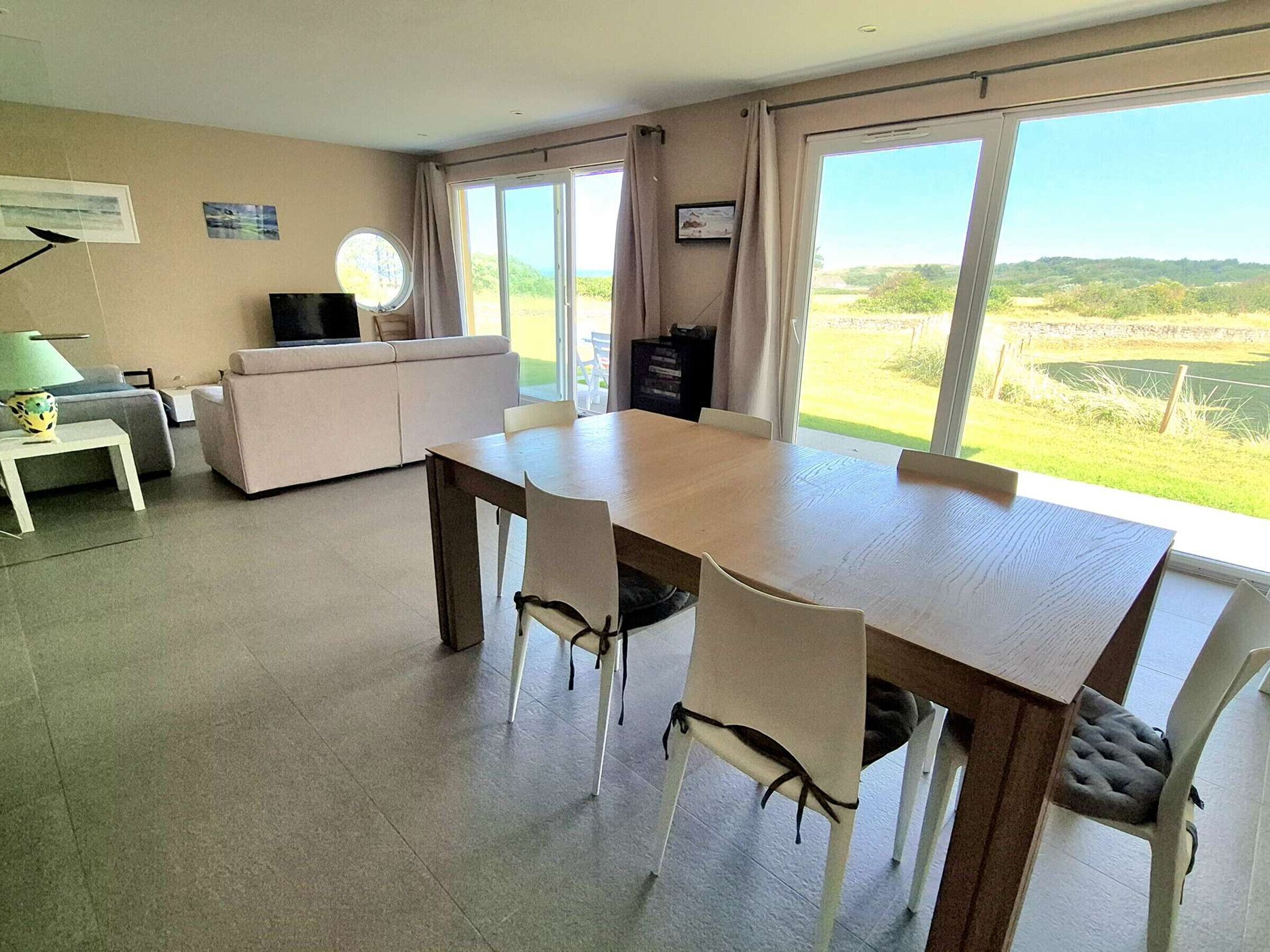 Een eethoek met een tafel en stoelen, een bank, een televisie en grote ramen met uitzicht op groen.