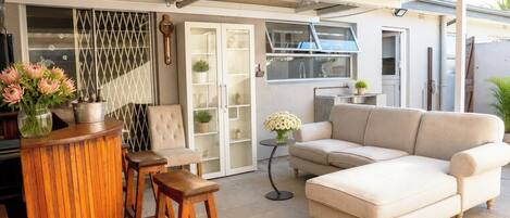 An outdoor seating area with a cushioned sofa, wooden stools, and a bar counter with a vase of flowers.