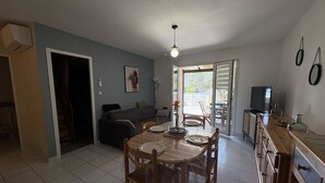 Un área de comedor con una mesa de madera, sillas y una lámpara en el techo. Se puede ver un sofá y un televisor al fondo.