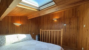 Un dormitorio con tragaluz, paredes de madera, una cama con ropa de cama estampada y dos lámparas fijadas a la pared.