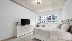 Un dormitorio con cama, televisión sobre una cómoda, ventana con vista a edificios y techo con vigas a la vista.