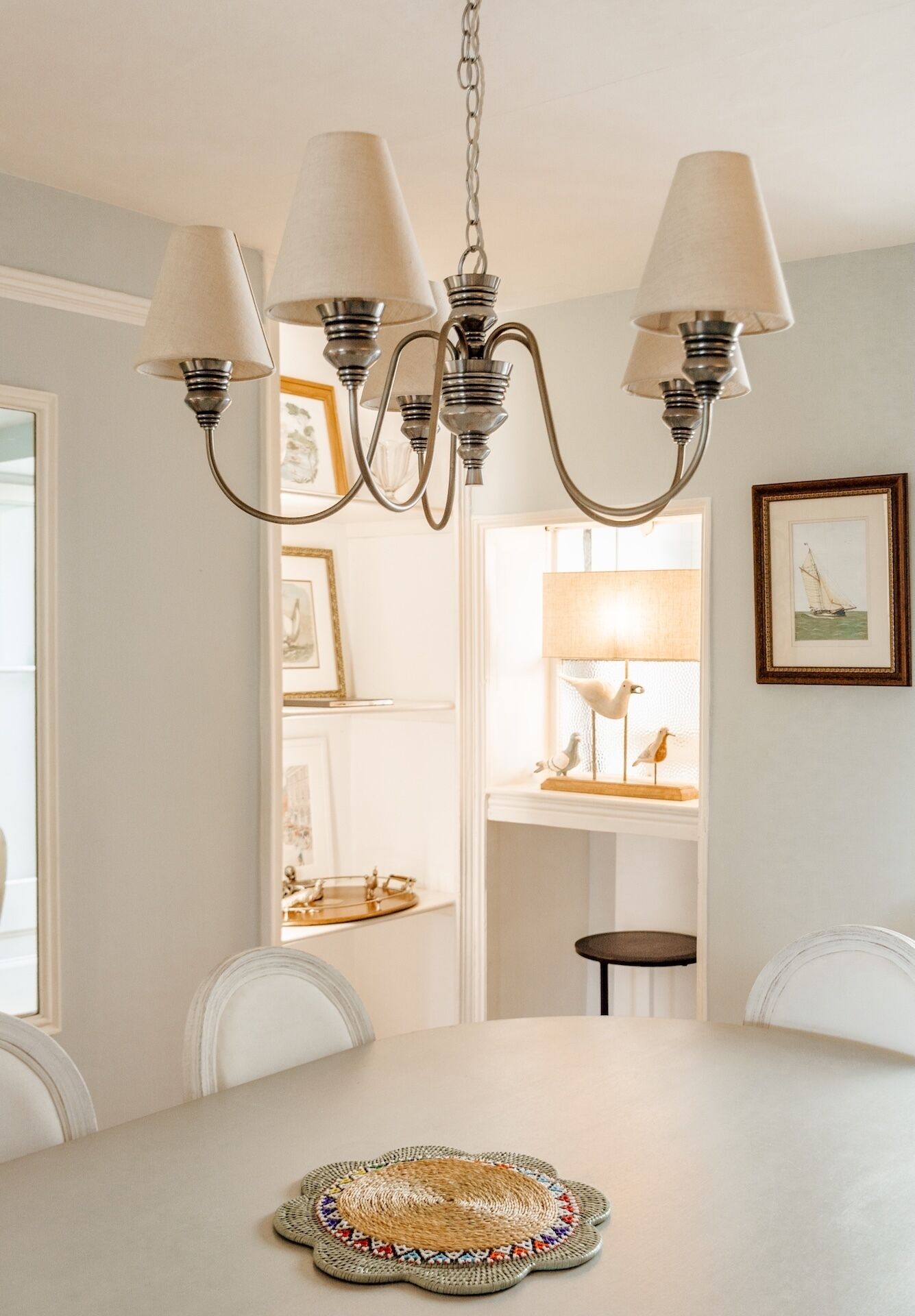 A dining area with a chandelier, white chairs, a table with a woven placemat, and framed artwork on the walls.