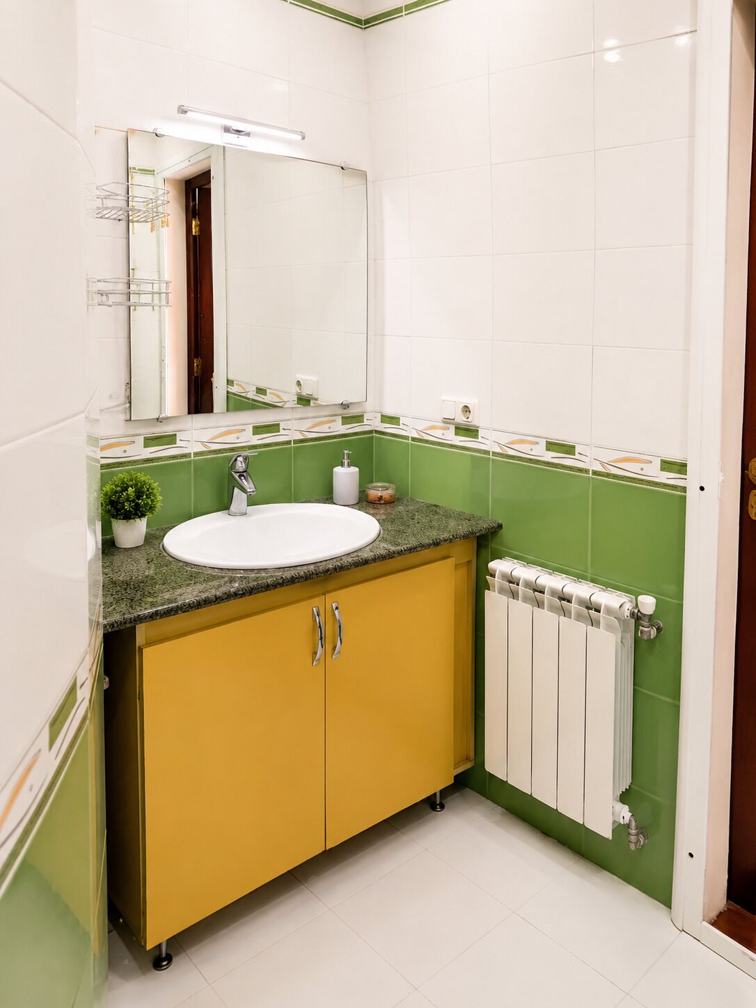 A bathroom with a white sink, green and white tiles, a yellow cabinet, a mirror, and a radiator.