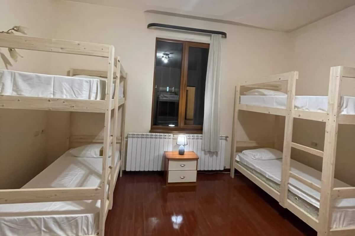 A room with two bunk beds, a small table with a lamp, a radiator, and a window with curtains.