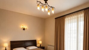A hotel room with a large bed, two bedside tables with lamps, a chandelier, and a large window with curtains.