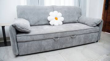 Un sofà gris amb un coixí amb forma de flor blanca, una taula petita i una porta de fusta al fons.