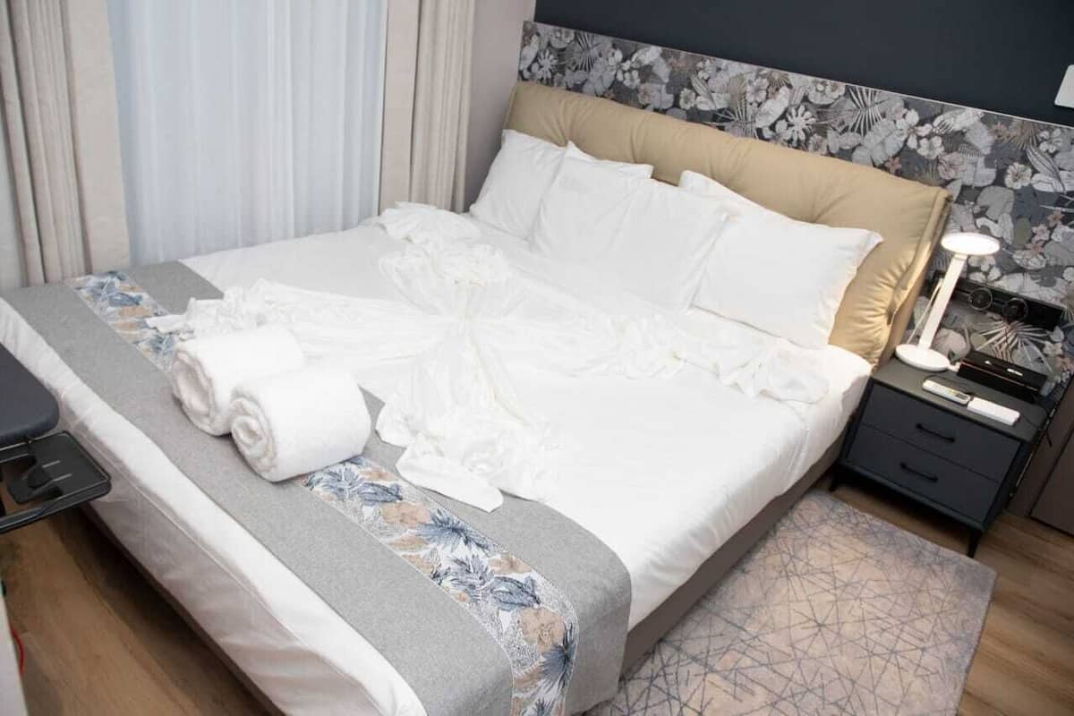 Una cama bien tendida con sábanas blancas, una mesita de noche con una lámpara y un cabecero estampado con motivos florales.