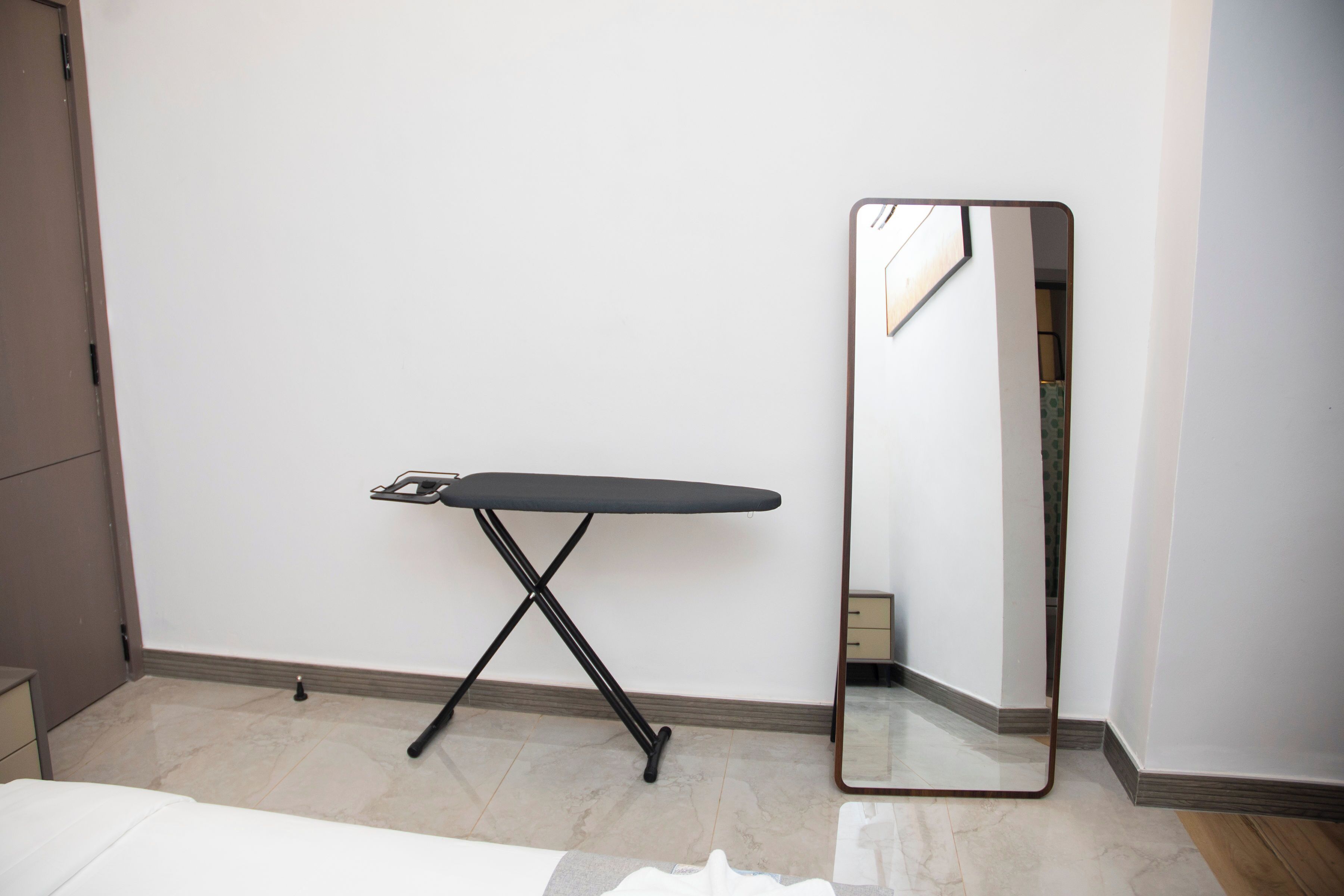 A bedroom with a bed, a small bedside table, a wall-mounted mirror, and a foldable ironing board.
