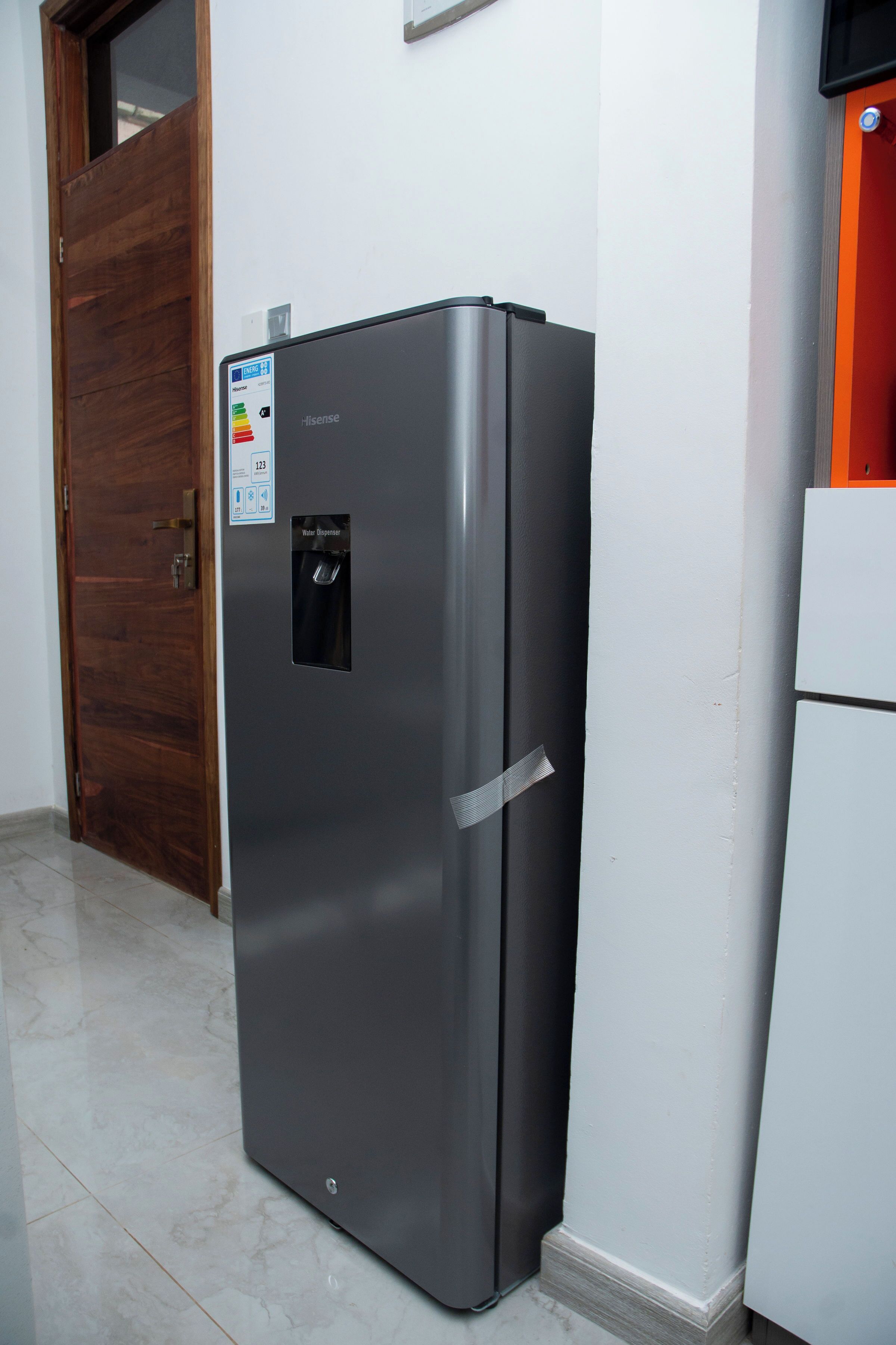 Ein kompakter, moderner Kühlschrank mit Wasserspender, platziert in einer Diele mit Holztür und weißer Wand.