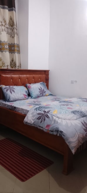 A single bed with a floral-patterned bedspread, a wooden headboard, and a red rug on the floor.