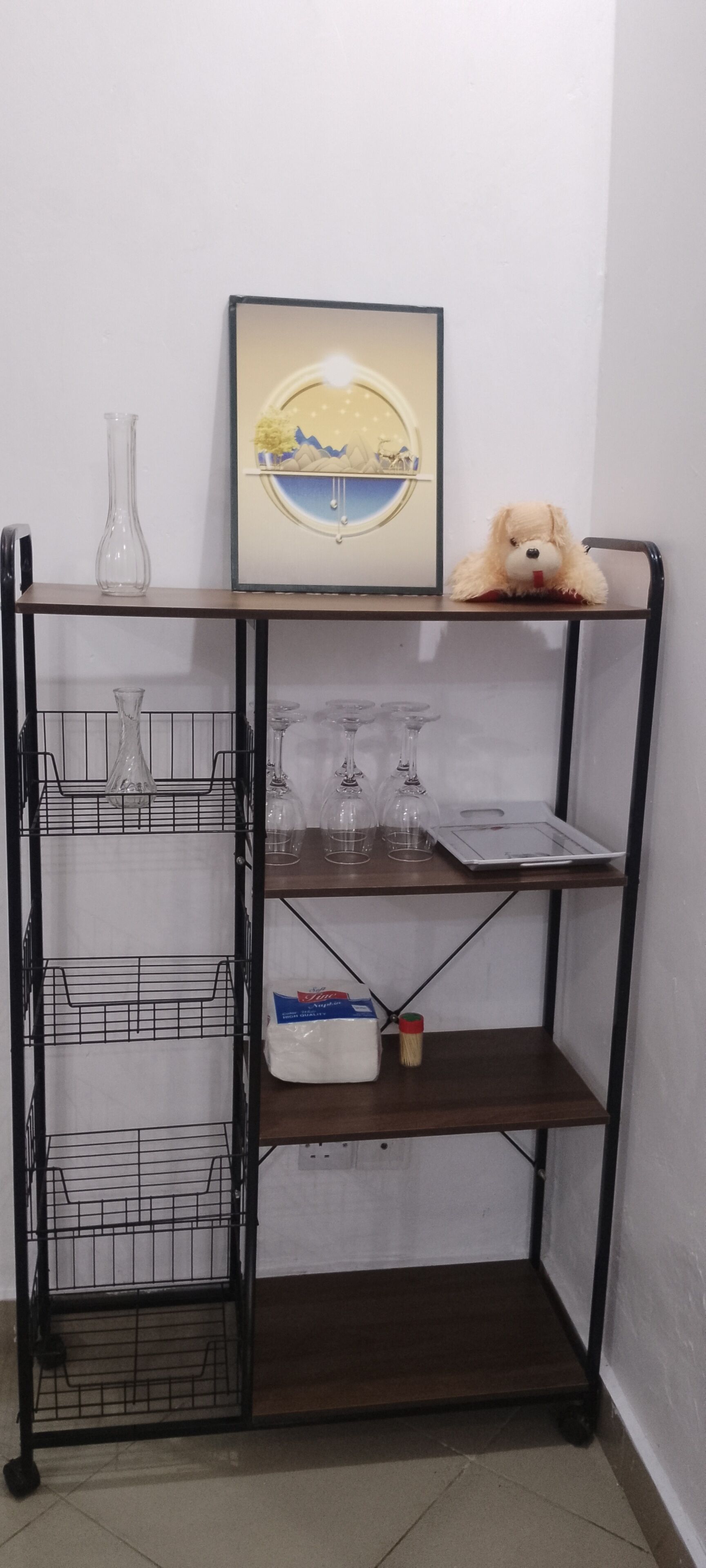 A metal shelf unit with multiple compartments, including a section with wine glasses and a section with a packaged item.
