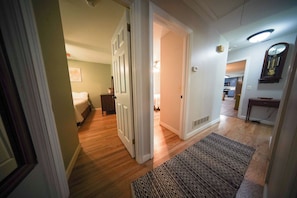 A hallway leading to a bedroom with a bed, a dresser, and a door. There is a rug on the floor and a clock on the wall.