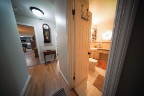A hallway leading to a bathroom with a toilet, sink, and mirror. There is a clock on the wall and a door with a towel rack.