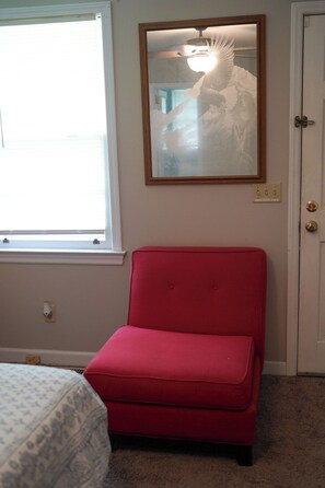 A room with a red chair, a bed with patterned bedding, a window with blinds, and a wall-mounted light fixture.