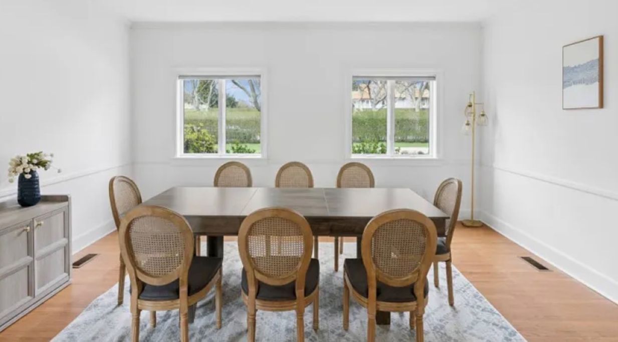 A dining room with a rectangular table, six wicker chairs, a grey cabinet, and two windows with a view of greenery.