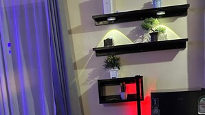 Een woonkamer met een bank, een zwarte koelkast, een potplant en een wandplank met een kleine plant en een waterfles.