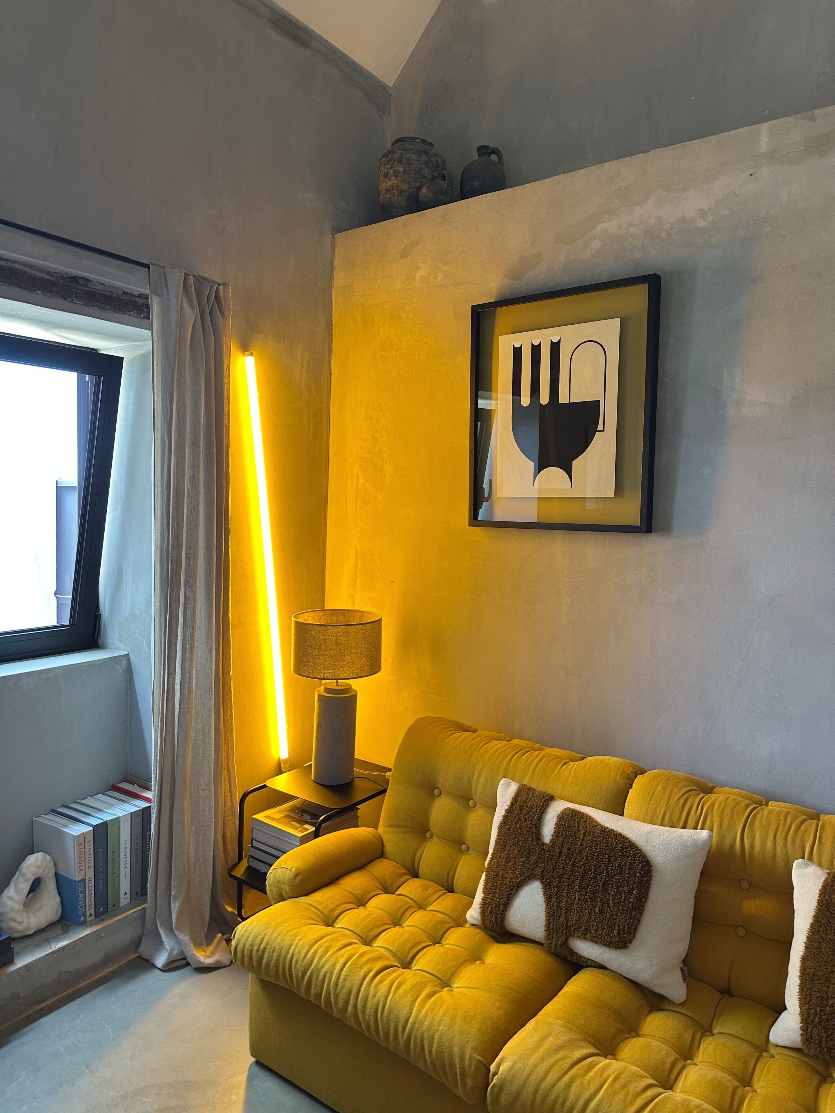 Una habitación con un sofá amarillo, una obra de arte enmarcada, una lámpara y una ventana con cortinas.