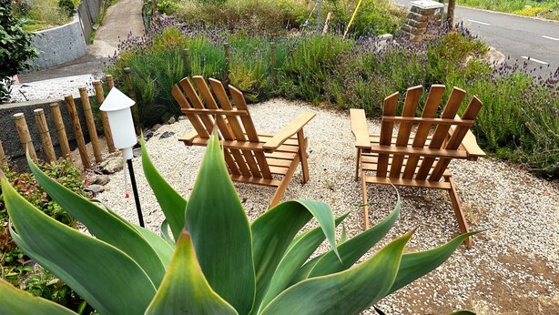 Two wooden chairs and a table set on a gravel surface with green plants in the foreground. - THE L Lodge, Ocean View Private Garden Cottage (Tabua)