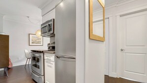 Une cuisine compacte dotée d’appareils électroménagers en acier inoxydable, d’un réfrigérateur blanc et d’un four à micro-ondes. Un tableau encadré orne le mur, et une porte au cadre doré est visible.