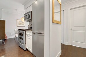 Eine kompakte Küche mit Edelstahlgeräten, einem weißen Kühlschrank und einer Mikrowelle. An der Wand hängt ein gerahmtes Bild und eine Tür besitzt einen goldenen Rahmen.