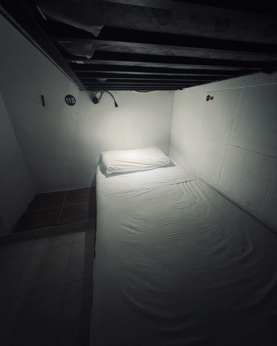 A small, dimly lit room with a single bed, a tiled floor, and a wall-mounted light fixture.