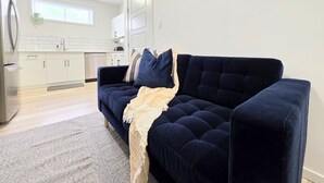 Sala de estar moderna com um sofá azul escuro, uma cozinha ao fundo e uma janela com persianas.